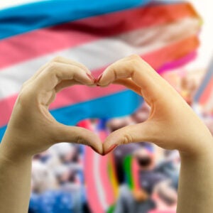 A young person's fingers make a heart shape, in front of the blue, lavender and white transgender rights flag.