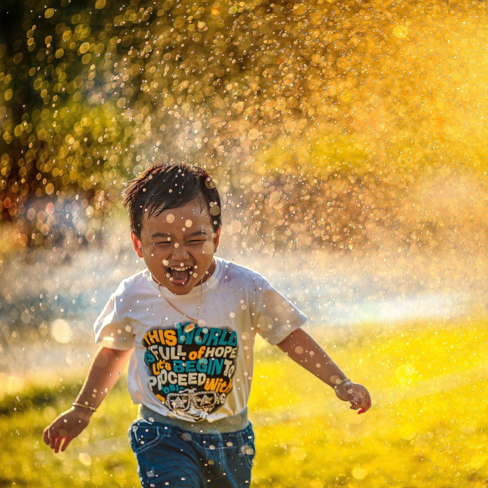 A young boy of Asian descent, joyfully running under a lawn sprinkler.