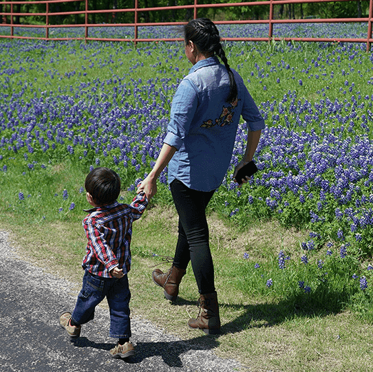 Mother and child holding hands while strolling past a lush field of bluebonnets.